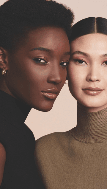 Duas mulheres maquiadas com sombra rosa sendo uma negra com brincos de argola uma mulher com cabelo escuro e tiara na cabeça