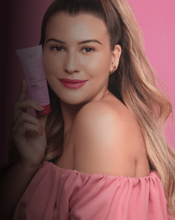 Niina Secrets, mulher branca de cabelos loiros utilizando batom de cor rosa e brinco dourado de argola na orelha esquerda, segurando um gel de limpeza da marca Eudora.