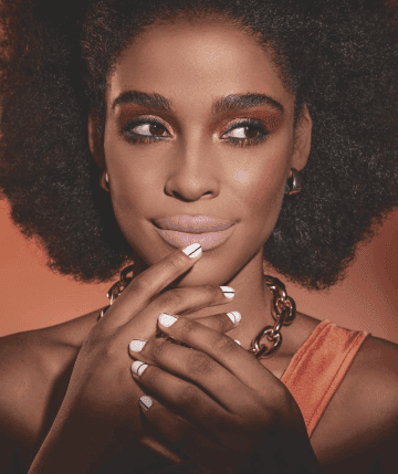Mulher negra maquiada com sombra vermelha, batom rosa, brincos de argola e colar, unhas brancas com linha preta e mão na boca.